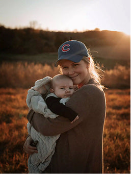 A smiling woman in a baseball cap holding a baby outdoors at sunset