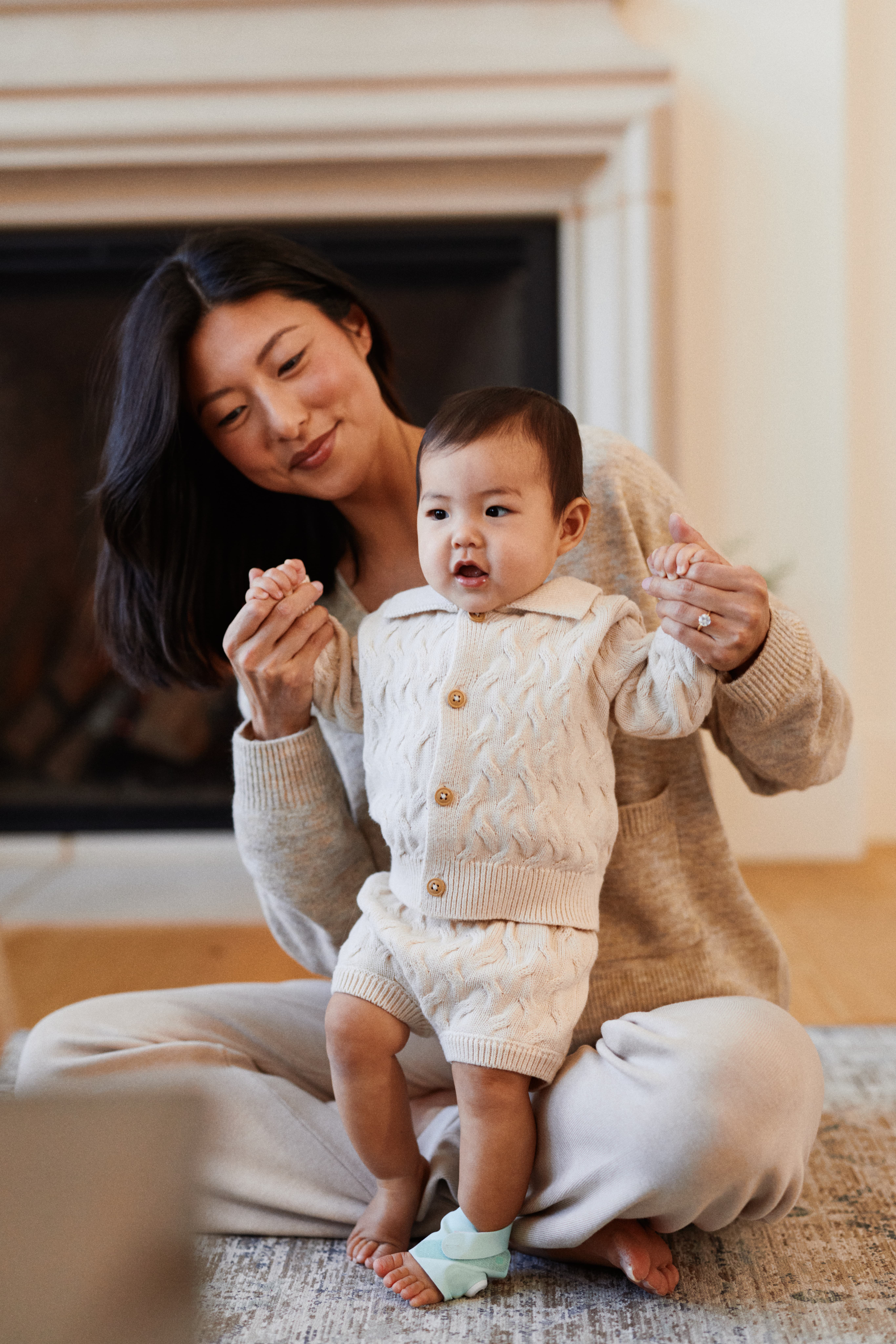 Parent helping their baby take first steps by the fireplace