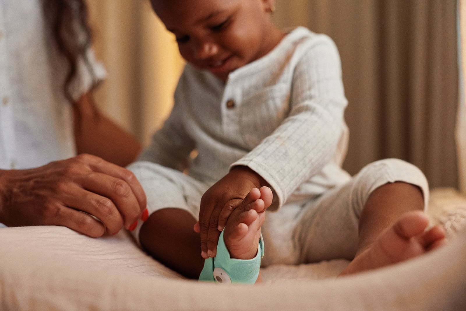 A toddler sitting on a bed pulling on their own Dream Sock, a parent beside them
